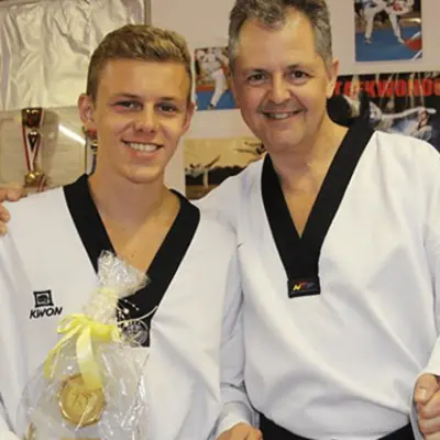 Zwei Taekwondoka im Dobok vor einer Wand mit Taekwondo-Fotos, der Jüngere hält einen Geschenkkorb