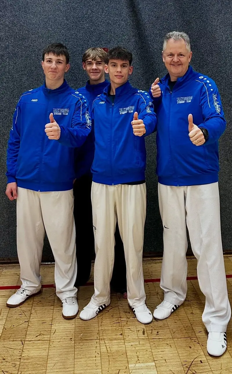 Drei Sportler und ihr Trainer in blauen Vereinsjacken, zwei der Sportler und der Trainer zeigen den Daumen nach oben