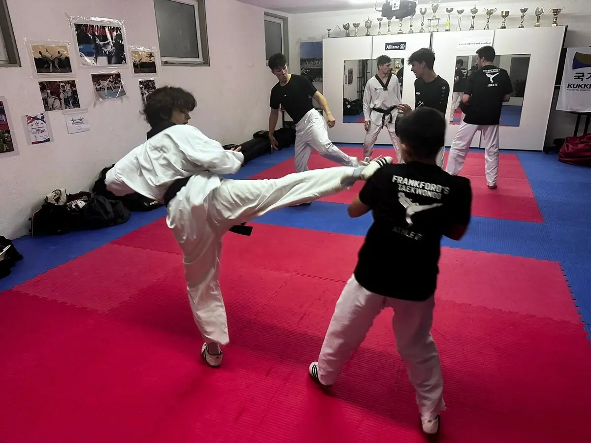 Partnertraining im Trainingsraum: Eine Person im Dobok mit schwarzem Gürtel tritt hoch, gegenüber ein Trainingspartner im Shirt von Frankford's Taekwondo